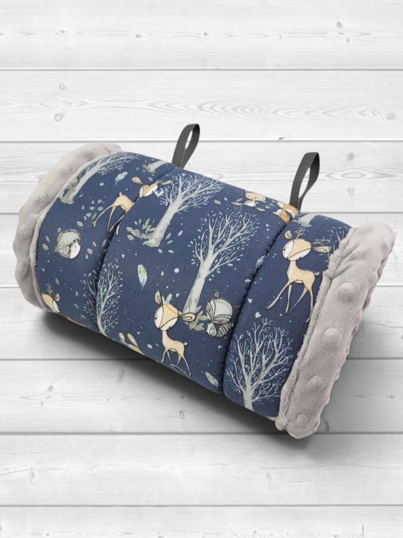 May include: A rolled-up fabric mat featuring a dark blue background with a repeating pattern of light brown deer, grey trees, and small animals. The ends of the mat are finished with a textured, light grey plush fabric and two black fabric loops.