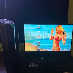 May include: A black laptop displaying a video game interface with a beach scene and a woman in a bikini. A black gaming console is visible to the left of the laptop.
