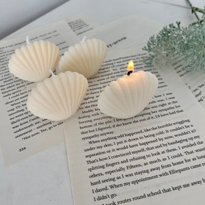 May include: A set of five white seashell-shaped candles. The candles are arranged in a semi-circle on a white surface. One of the candles is lit and has a small flame.