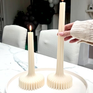 May include: Two ivory-coloured, lit taper candles with a fluted base sit on a white tray. One candle is held by a person. The candles are on a white marble table. The background includes white chairs and black and white balloons.
