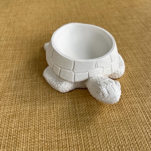 May include: A white turtle-shaped planter with a round opening. The turtle has a textured shell and head, and is sitting on a textured surface. The planter is made of a solid material.