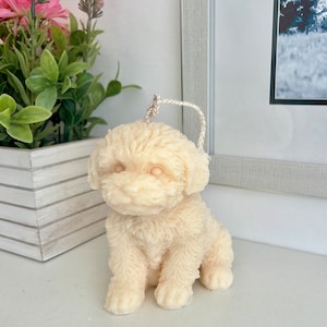May include: A white, fluffy, dog-shaped candle with a cream-coloured string loop on top. The candle is sitting on a white surface.