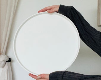 May include: A white round tray being held by a person's hands. The tray is made of a smooth, solid material.