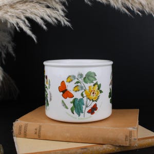 May include: A white ceramic cylindrical container featuring a botanical illustration of a yellow Gossypium barbadense flower with green leaves and two orange butterflies. The text Gossypium barbadense is printed in small green letters near the base.
