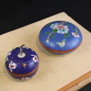 May include: Two cloisonné enamel containers featuring a deep blue background with intricate gold wire patterns, floral motifs in pink and white, and green leaves. One container is apple-shaped with a small metal stem, while the other is round.