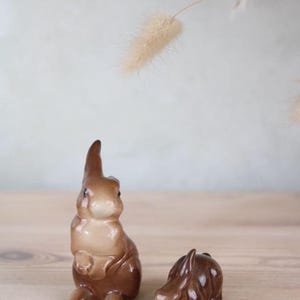 May include: Two brown ceramic bunny figurines. One is upright, the other is lying down. They are on a wooden surface, with a dried plant in the background. The bunnies are decorative and likely for Easter.