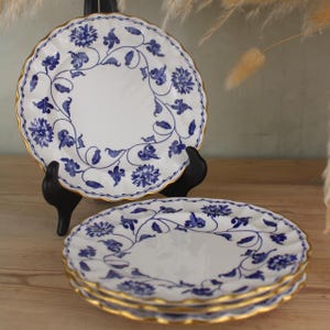 May include: White porcelain plates with a blue floral pattern and gold trim. The plates have a scalloped edge and are displayed on a wooden surface. The plates are stacked and displayed on a black stand.