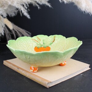Peut inclure: Un bol en céramique en forme de feuille de chou avec un extérieur et un intérieur vert clair. Le centre présente un motif de fruit orange peint avec des feuilles vertes. Le bol est soutenu par trois petits pieds ronds à pois orange.