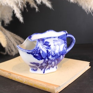 May include: A white ceramic shaving scuttle featuring a blue floral pattern with a scalloped rim and a side handle. The vessel has a distinct interior shelf for holding soap and a wide opening for lathering, decorated with detailed blue rose motifs.
