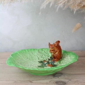 Peut inclure: Un plat de service en céramique en forme de feuille de chou, avec un glaçage vert vif. Une figurine d'écureuil marron est au centre, tenant une noix. Le plat mesure environ 30 cm de diamètre.