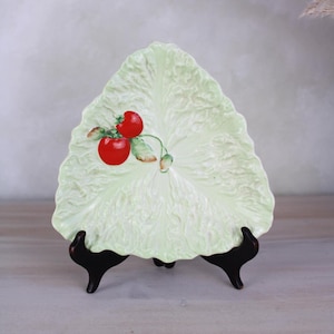 Peut inclure: Un plat de service en céramique vert en forme de feuille de laitue avec deux tomates rouges peintes dessus. Le plat est sur un support noir.