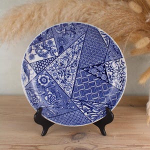 May include: A round, blue and white decorative plate with a patchwork design. The plate features various floral and geometric patterns in shades of blue. It is displayed on a black stand, showcasing its intricate details and craftsmanship. The plate is a decorative item.