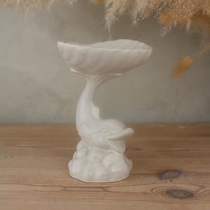 Peut inclure: Un plat en céramique blanche en forme de dauphin. Le plat a un bord festonné et est soutenu par une figurine de dauphin s'élevant d'une base en forme de nuage. Le dauphin sourit. Le plat est sur une surface en bois.
