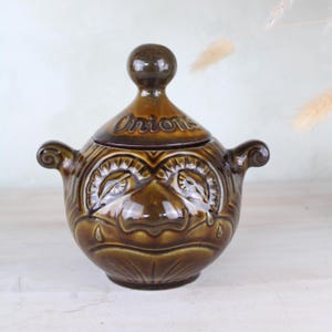 May include: A brown ceramic onion pot with a lid. The lid is inscribed with the word "Onions". The pot has two handles and a decorative face design. The ceramic has a glossy finish.