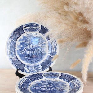 May include: Blue and white decorative plates with a nautical theme. The plates feature a detailed illustration of a sailing ship on the ocean. The plates have a patterned border and are stacked on a wooden surface.