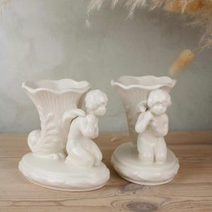 Peut inclure: Deux vases en céramique blanc cassé, chacun orné d'un chérubin agenouillé avec des ailes. Les vases ont une forme de trompette cannelée avec des détails en relief décoratifs. Les chérubins sont agenouillés sur une base ovale.