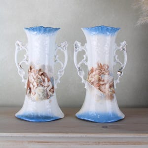 Peut inclure: Paire de vases en porcelaine vintage avec des accents bleus et blancs. Chaque vase présente un motif peint à la main de chérubins jouant de la musique.