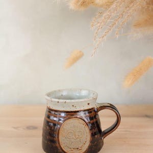 May include: A handmade ceramic mug with a brown and cream color scheme. The mug features a circular emblem with a design and a dark brown handle. The mug is sitting on a wooden surface.
