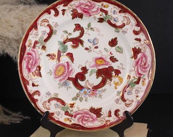 May include: A round ceramic plate featuring a white center with a floral pattern of pink peonies, green leaves, and dark red scrollwork. The scalloped rim is decorated with a wide dark red border, gold accents, and additional pink floral motifs.