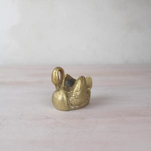 May include: A small, brass swan ornament with intricate detailing. The swan features a curved neck and a small, open bowl-like space on its back. The ornament is a warm, golden hue and rests on a pale pink surface.