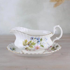 Peut inclure: Un saucier en porcelaine blanche avec un motif floral dans les tons de bleu, rose et vert. Le saucier a une bordure dorée et repose sur une soucoupe assortie.