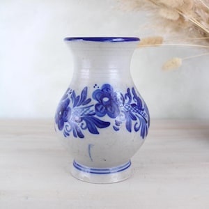 Könnte beinhalten: Eine kleine, weiße Keramikvase mit blauem Blumendesign. Die Vase hat einen runden Körper und einen ausgestellten Rand.