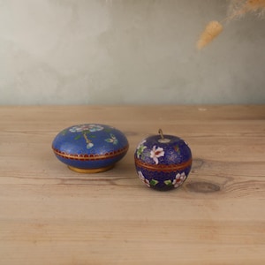 Könnte beinhalten: Zwei blaue Cloisonné-Schmuckkästchen. Eines ist rund mit einem Blumendesign und einem goldenen Sockel. Das andere ist apfelförmig mit einem Blumenmuster und einem kleinen Metallstiel. Beide haben aufwendige Details und eine satte Farbpalette.