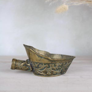 May include: A small, antique brass bowl with an ornate floral design. The bowl has a spout on one side and is perfect for holding small items.