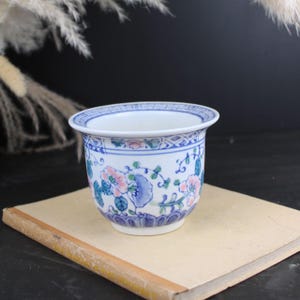 May include: A white ceramic planter featuring a blue and pink floral pattern with green leaves. The rim is decorated with a blue geometric border, and the base is accented with a blue petal design. The vessel has a wide, flared opening.