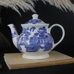 May include: A white ceramic teapot with a blue Willow pattern design. The teapot has a curved spout, a rounded handle, and a lid with a small knob. The blue and white design depicts a landscape scene with buildings, trees, and a bridge. The teapot is on a tan surface.