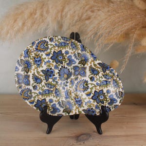 May include: An oval-shaped ceramic plate with a white background and a blue floral pattern. The plate is supported by a black stand, and the design includes yellow and green accents. The plate is displayed on a wooden surface.