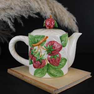 Könnte beinhalten: Eine cremefarbene Keramik-Teekanne mit einem erhabenen, handbemalten Erdbeermotiv mit grünen Blättern und roten Beeren. Der Deckel ist mit einem kleinen Erdbeerknauf versehen und der Henkel weist eine gedrehte, seilartige Struktur auf.