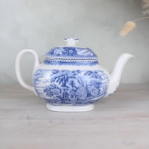May include: A white ceramic teapot with a blue landscape design. The teapot features a lid with a floral pattern and a curved handle. The design includes a house, trees, and a river. The teapot is a classic design.