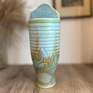 Peut inclure: Un vase en céramique bleu clair avec un glaçage vert et brun. Le vase a un design unique avec un sommet festonné et un motif décoratif de feuilles et de volutes.