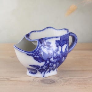 Könnte beinhalten: Eine weiß-blaue Rasierkelle aus Keramik mit floralem Design. Die Kelle hat einen Griff und eine einzigartige Form mit gewelltem Rand und einem separaten Fach für Seife. Das blaue Blumenmuster bedeckt den größten Teil der Oberfläche.