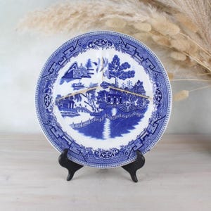 Könnte beinhalten: Ein blauer und weißer Porzellanteller mit einem Weidenmuster. Der Teller ist gerissen und hat einen kleinen Absplitterung am Rand. Er wird auf einem schwarzen Ständer präsentiert.
