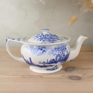 May include: A white ceramic teapot with a blue landscape design. The teapot has a curved handle and spout, and a lid with a matching design. The design features trees, buildings, and other landscape elements. The teapot is sitting on a wooden surface.