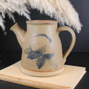 May include: A tan stoneware jug with a speckled texture and a blue painted leaf design on the front. The jug features a wide mouth, a single side handle, and a short spout. The surface shows horizontal ridges around the body.