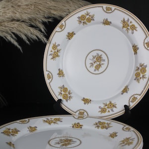 Peut inclure: Assiettes en porcelaine blanche avec un motif floral jaune et une bordure dorée. Les assiettes présentent un médaillon floral central et des motifs floraux répétés sur le bord. Les assiettes sont empilées et présentées sur fond noir.