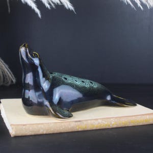 May include: A ceramic seal ornament in dark blue and green with a glossy finish. The seal is lying down with its head raised. The figurine has a series of small holes along its back. The seal is resting on a book.