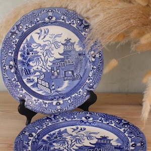 May include: Two blue and white decorative plates with a classic Willow pattern. The plates feature a detailed landscape scene with pagodas, bridges, and figures. The plates are displayed on black stands, showcasing their intricate design.