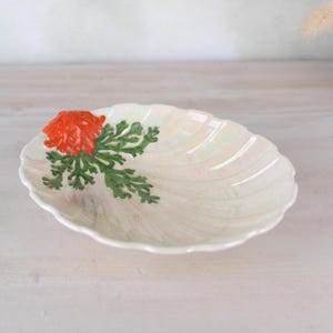 May include: A white ceramic dish with a pearlescent finish shaped like a seashell. The dish has a raised orange and green flower design.