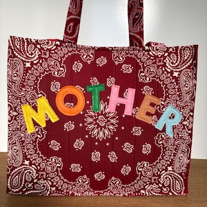May include: A quilted tote bag made from dark red fabric with a white paisley pattern. The bag features two handles and the word MOTHER appliqued across the front in large, colorful felt letters: yellow M, orange O, green T, pink H, brown E, and light blue R.
