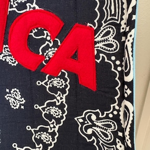 May include: A quilted fabric item featuring a dark navy blue and white paisley pattern. Large, bold, red felt letters spelling CA are appliqued onto the surface. The edge of the fabric is finished with a light blue trim, and vertical stitching lines run across the quilted material.