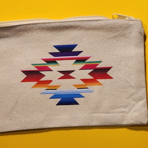 May include: A beige canvas zippered pouch with a colorful geometric design printed on the front. The design features a repeating pattern of triangles and stripes in shades of red, orange, yellow, green, blue, purple, and pink.