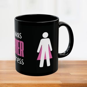 May include: Black ceramic mug with the text "I was NEVER a waitress" in white and pink. A white figure with a pink cape is also on the mug. The mug is on a wooden surface.