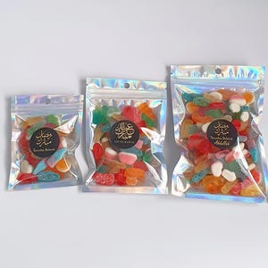 May include: Three clear plastic bags with zipper closures filled with colorful gummy candies. The bags are different sizes and have a holographic iridescent finish. The bags have labels with Arabic text and the words "Ramadan Mubarak" and "Eid Mubarak".