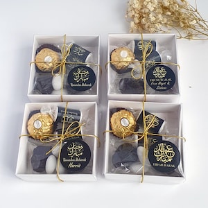 May include: Four white gift boxes with gold ribbon and clear cellophane windows. Each box contains a chocolate, a small white candy, and a black and gold sticker with Arabic calligraphy and the text "Ramadan Mubarak" or "Eid Mubarak".