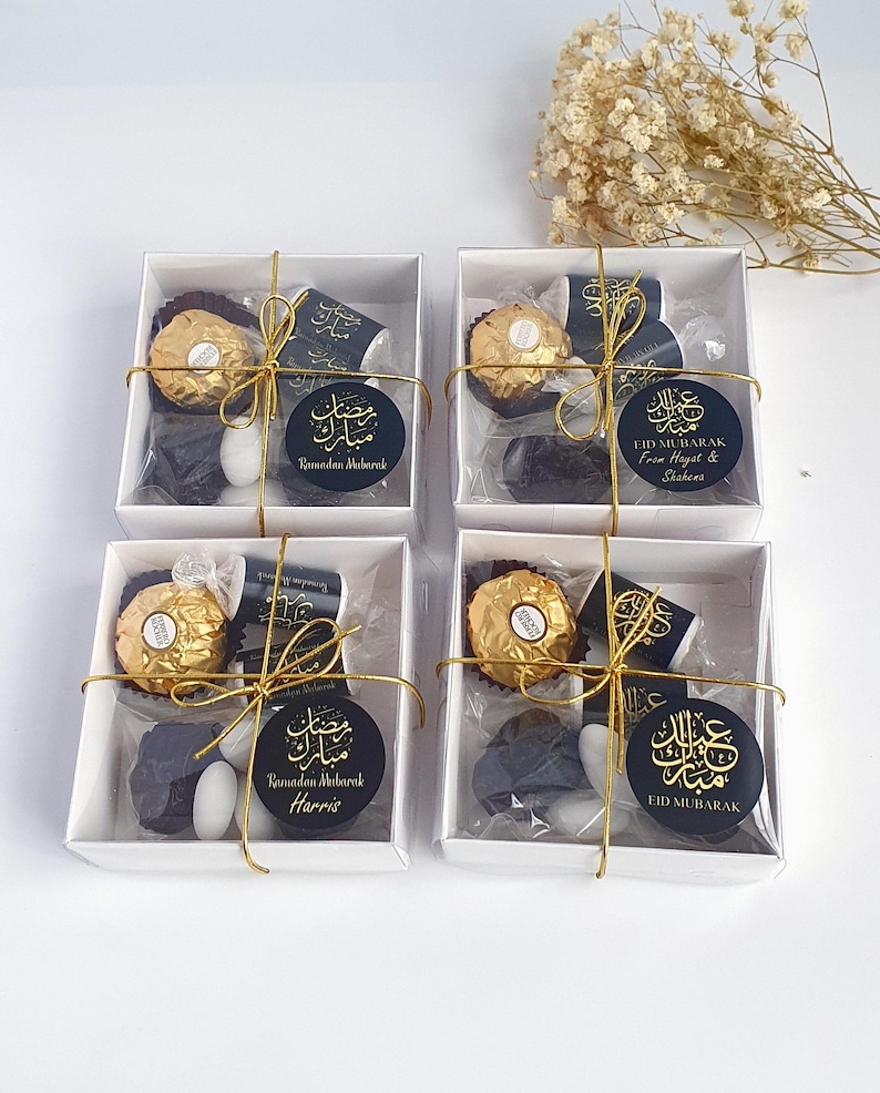 May include: Four white gift boxes with gold ribbon and clear cellophane windows. Each box contains a chocolate, a small white candy, and a black and gold sticker with Arabic calligraphy and the text "Ramadan Mubarak" or "Eid Mubarak".
