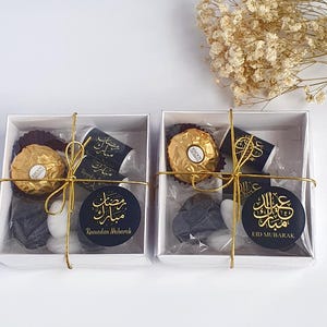May include: Two white gift boxes with gold ribbon and black and gold labels. The boxes contain chocolate, candy, and a small black gift. The labels on the boxes say "Ramadan Mubarak" and "Eid Mubarak".